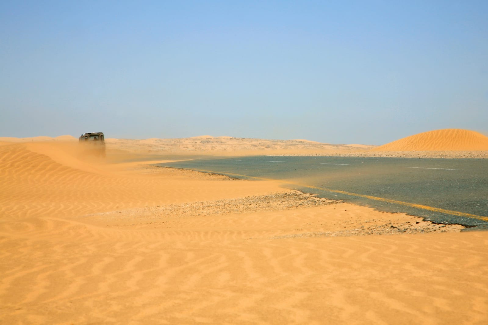 Sand weht in flachen Streifen über eine Asphaltstraße, am Rand liegen Dünen