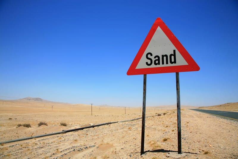 Warnschild mit der Aufschrift Sand neben einer Straße, dahinter eine versandete Bahnlinie