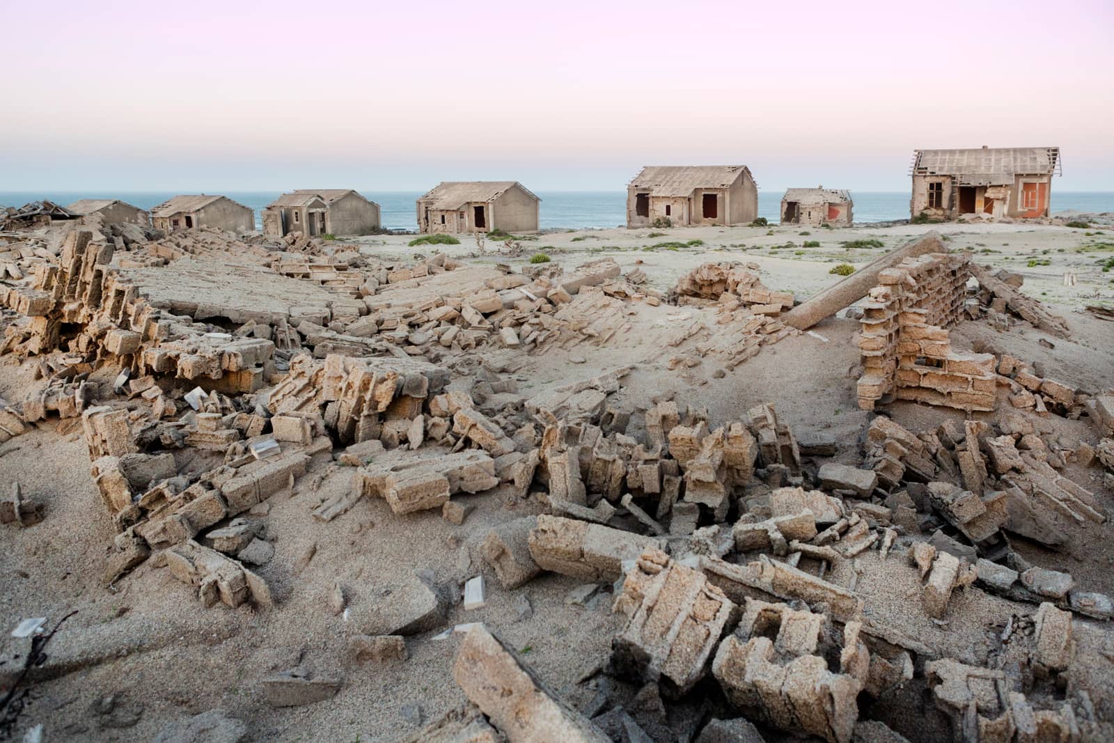 Eingestürzte Mauern und verlassene Häuser im Sand, dahinter das Meer