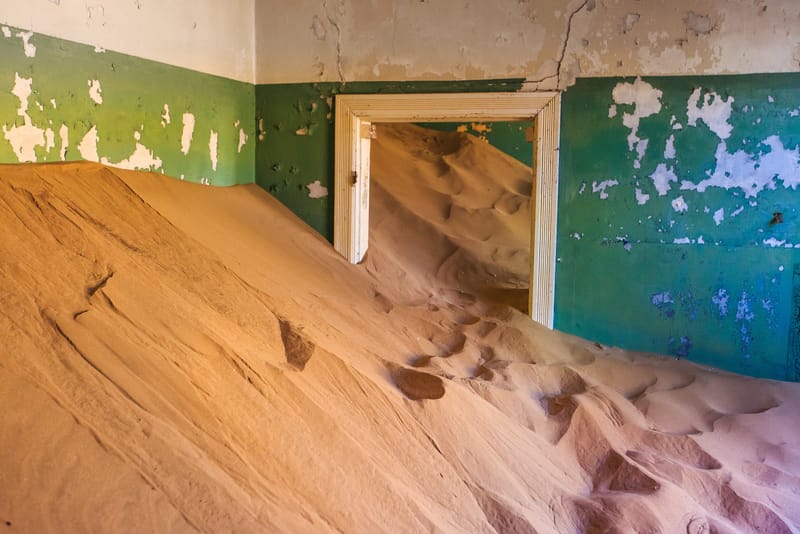 Raum mit abblätternder grüner Farbe, durch die Tür fließt eine Sanddüne herein